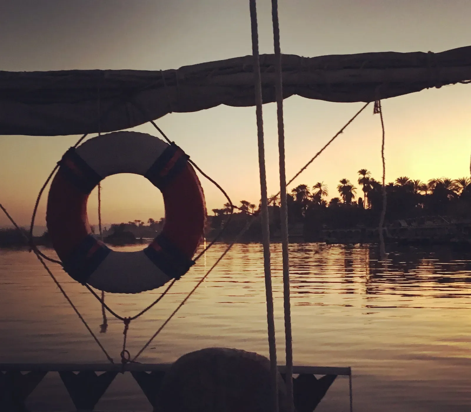 A life preserver ring hangs from a felucca's rigging, framing a view of silhouetted palm trees and the Nile at sunset.