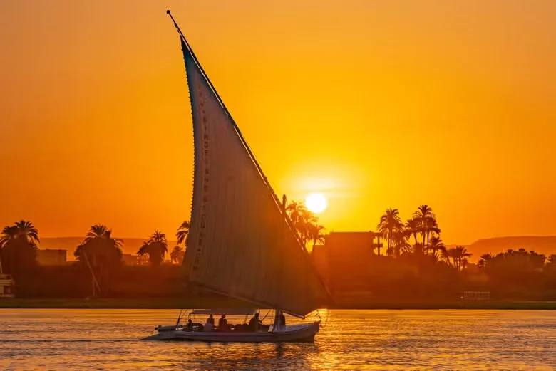 Fehér felucca-vitorla sziluettje narancssárga naplemente előtt pálmákkal a Nílus partján