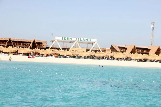 منظر واسع من الماء لأكواخ الشاطئ بسقف القش ولافتة جزيرة إيدن، مع كراسي استلقاء على رمال بيضاء