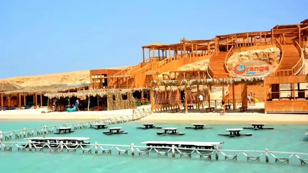 The wooden Orange Bay resort building on a sandy hillside with its logo sign and submerged picnic tables in the sea.