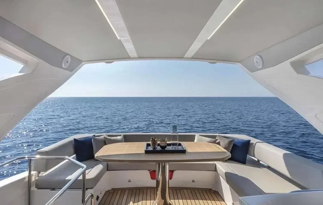 Rear lounge deck of a modern motor yacht with a curved white sofa and wooden table, open sea visible behind.