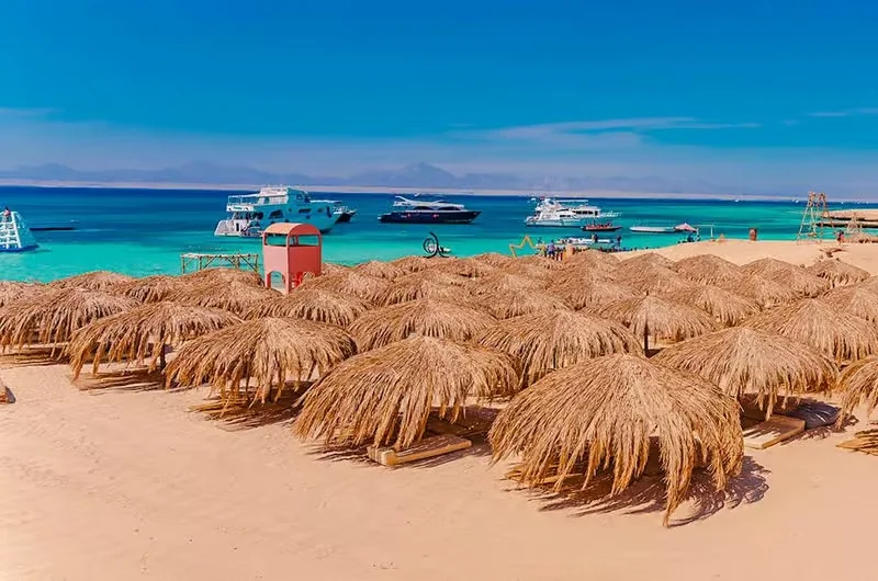Rieten strandparasols op goudgeel zand met enkele excursieboten voor anker in de turquoise baai en wazige bergen erachter