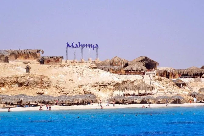 A Mahmya tábla homokos sziklák és nádtetős strandkunyhók fölé emelkedik, a türkizkék vízről nézve, alul strandolókkal