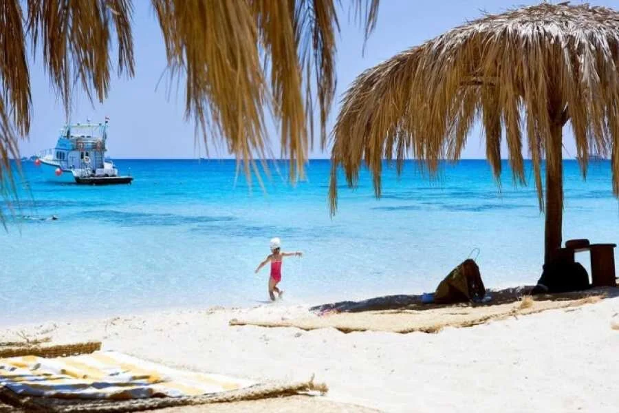 A Mahmya tábla és nádtetős kunyhók egy homokos sziklán egy zsúfolt strand felett, elhaladó hajóról nézve