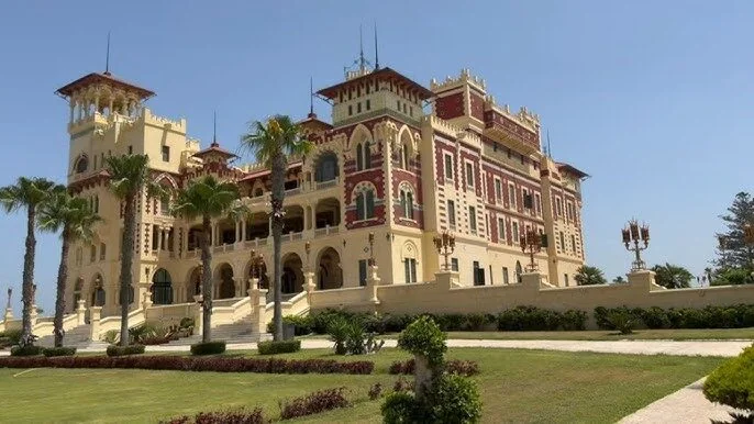 Ground-level view of the palace's arched facade and clock tower framed by tall palm trees on a sunny day.