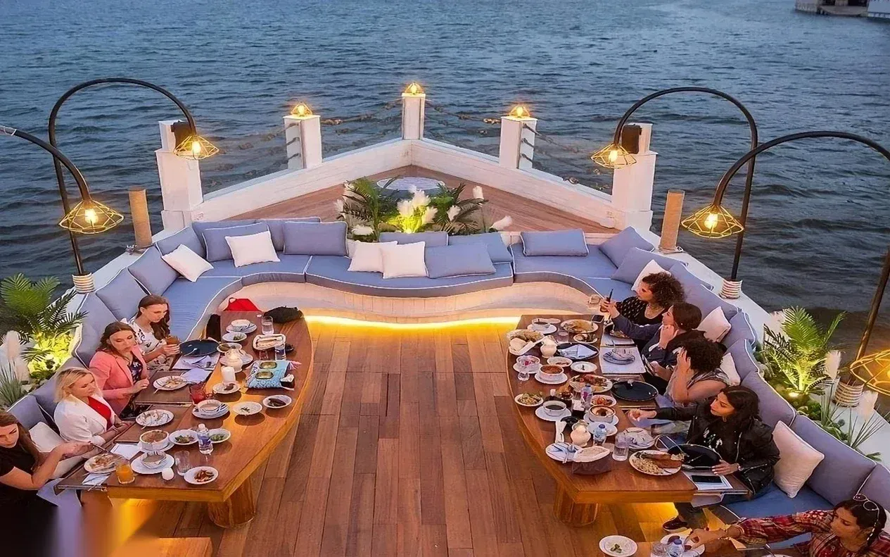 A similar aerial view of the same bow dining area, with guests eating and river lights reflecting on the water.