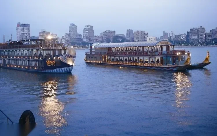 Two illuminated Nile dinner-cruise boats pass each other on the river at dusk, with the Cairo skyline behind them.