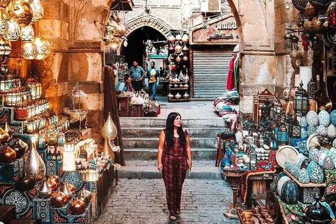 A woman in a patterned jumpsuit walks through a Khan el-Khalili alley lined with hanging lanterns, brass ornaments