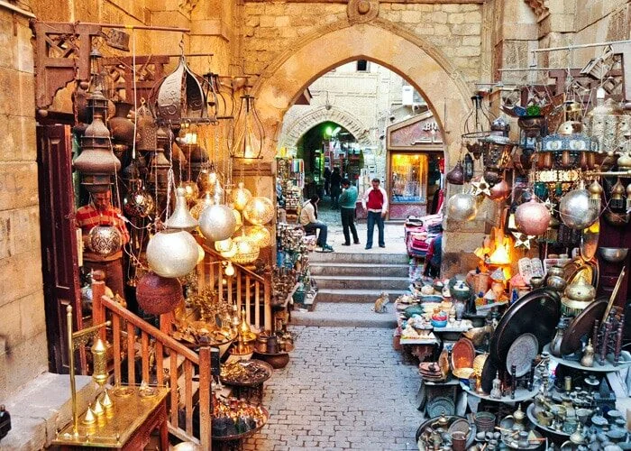 A narrow lantern-lined alley in Khan el-Khalili leads through a stone archway toward more market stalls