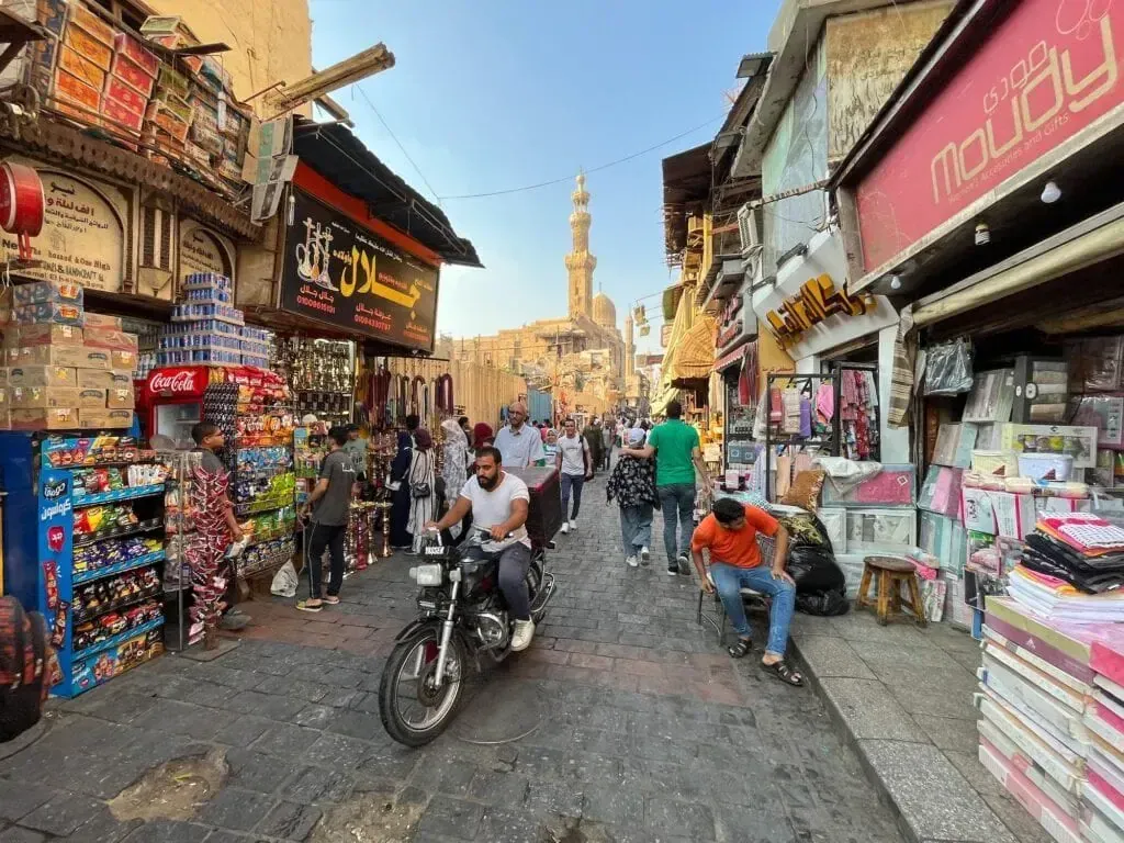 A busy Cairo market street lined with shops and stalls, with a motorbike rider and pedestrians passing beneath a distant