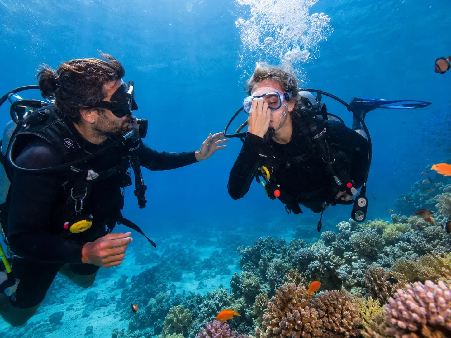 Instructor reaching toward a student diver who is adjusting her mask above a coral reef with small reef fish nearby.