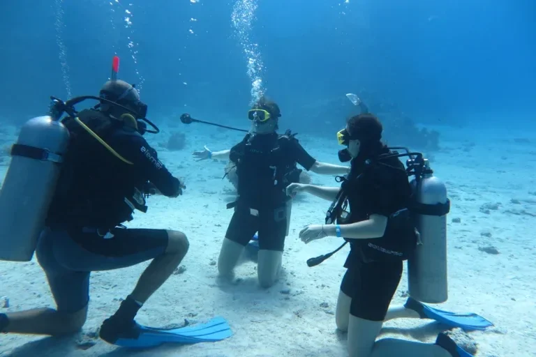 Three divers kneeling on a sandy seabed practicing skills, one diver facing away from the camera in the foreground.
