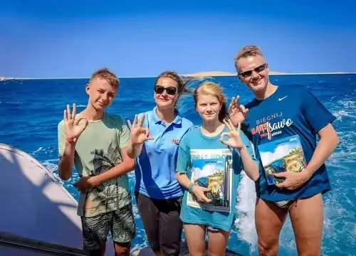 Vierköpfige Familie auf einem Tauchboot zeigt das OK-Zeichen, zwei halten Reisemagazine, im Hintergrund eine Wüsteninsel