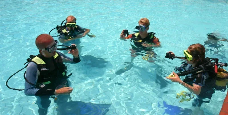 Vier junge Taucher knien im flachen Schwimmbecken und üben grundlegende Tauchfertigkeiten