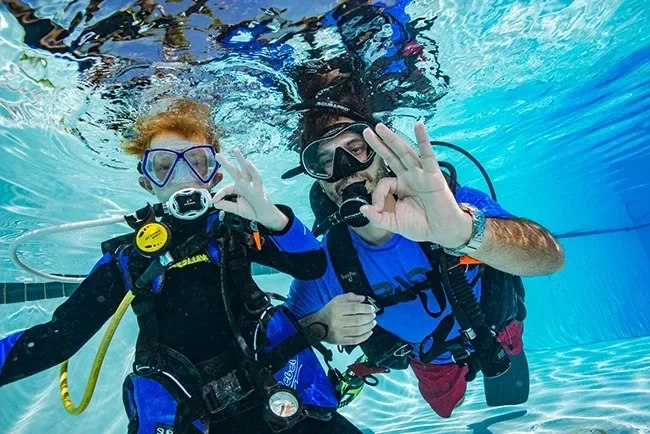 Zwei Taucher, einer ein rothaariges Kind, zeigen gemeinsam das OK-Zeichen unter Wasser im Schwimmbecken