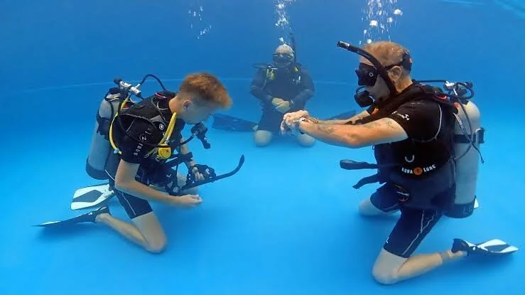 Zwei junge Taucher knien sich bei einer Übung im Schwimmbecken gegenüber, im Hintergrund ein Ausbilder