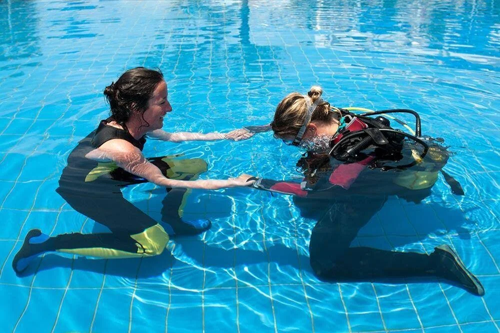 Instructor supporting a student diver as she practices mask and regulator skills at the surface of a swimming pool.