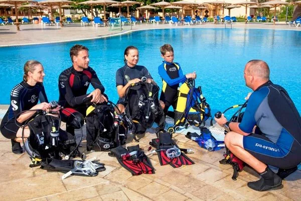 Group of four students and an instructor sitting poolside with dive gear laid out in front of them