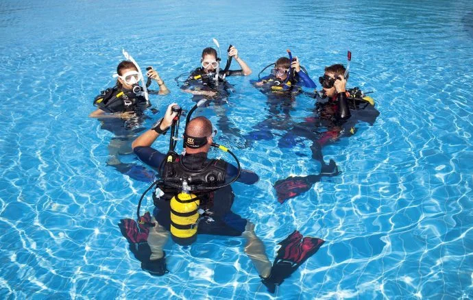 Instructor kneeling on a sandy seabed giving briefing hand signals to three student divers gathered around him underwater.
