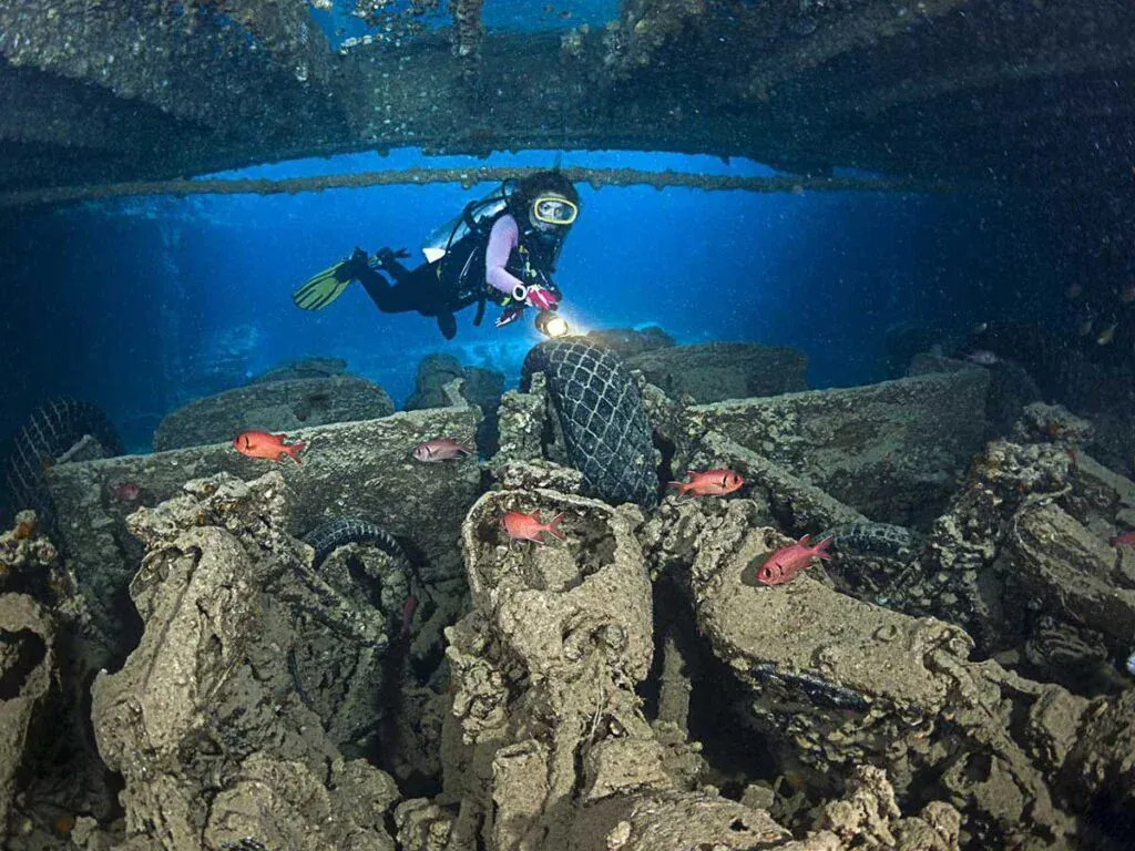 Taucher leuchtet mit der Lampe auf Trümmer und alte Reifen auf dem Deck eines Wracks, kleine rote Fische in der Nähe