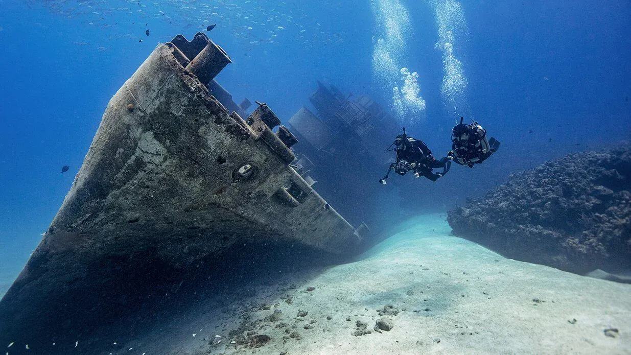 Zwei Taucher nähern sich dem hoch aufragenden Bug eines großen gesunkenen Schiffes, umgeben von Fischschwärmen