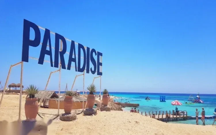 Groot blauw PARADISE-bord op het strand boven rieten cabana's en een druk turquoise zwemgebied