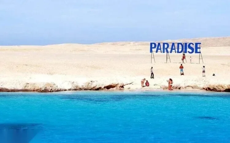 Toeristen staan aan de waterkant op een zandduin onder een PARADISE-bord, turquoise water op de voorgrond