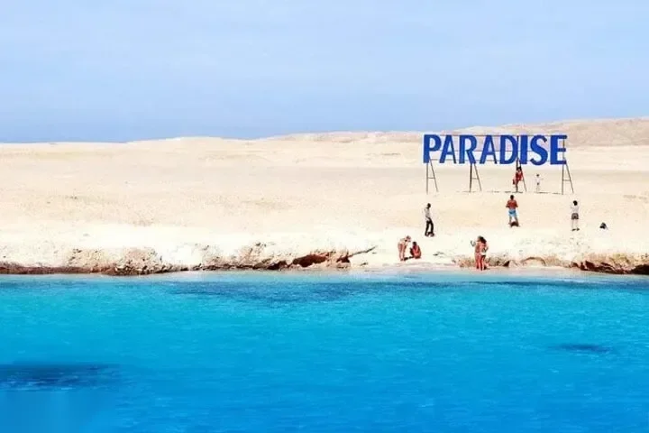 Toeristen staan aan de waterkant op een zandduin onder een PARADISE-bord, turquoise water op de voorgrond