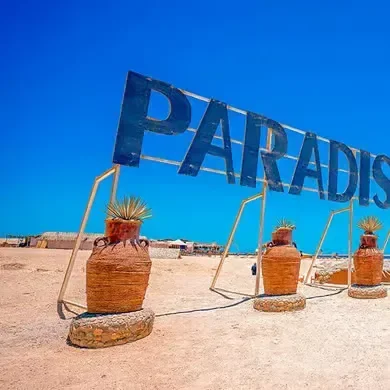 Een houten PARADISE-bord tussen terracotta potten op het zand onder een heldere hemel