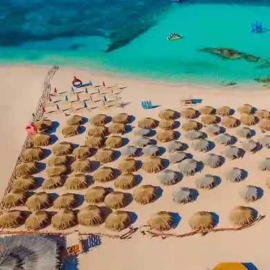 Luchtfoto van rijen rieten strandparasols op wit zand naast een turquoise baai