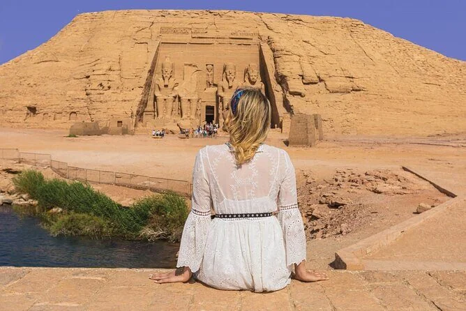 Frau in weißem Spitzenkleid sitzt mit dem Rücken zur Kamera und blickt über das Wasser auf den Großen Tempel von Abu Simbel