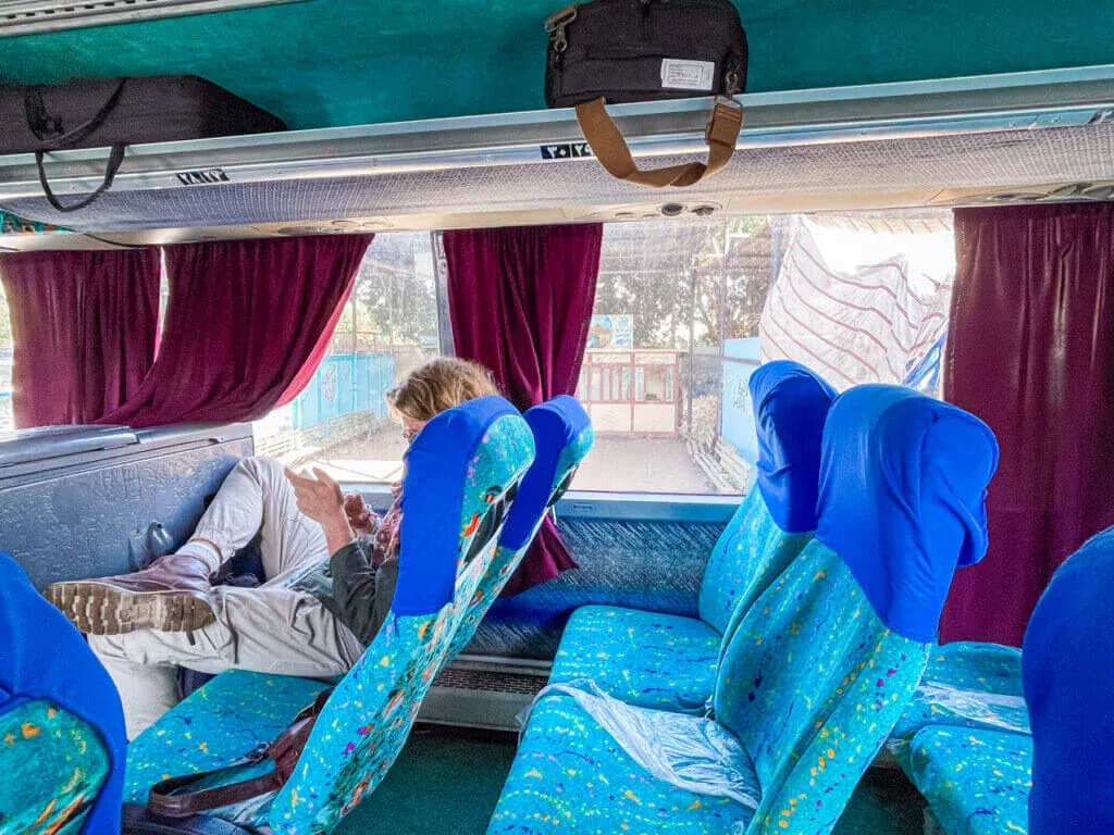 Reisebus von innen, ein Passagier liest ausgestreckt auf blauen Sitzen, bordeauxrote Vorhänge am Fenster