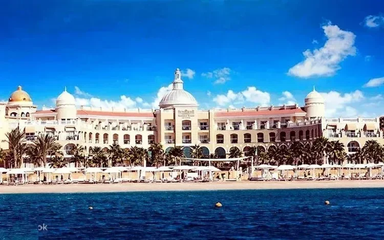 Large white resort hotel with domed towers along the Alexandria coastline, sun loungers and umbrellas lining the beach below.