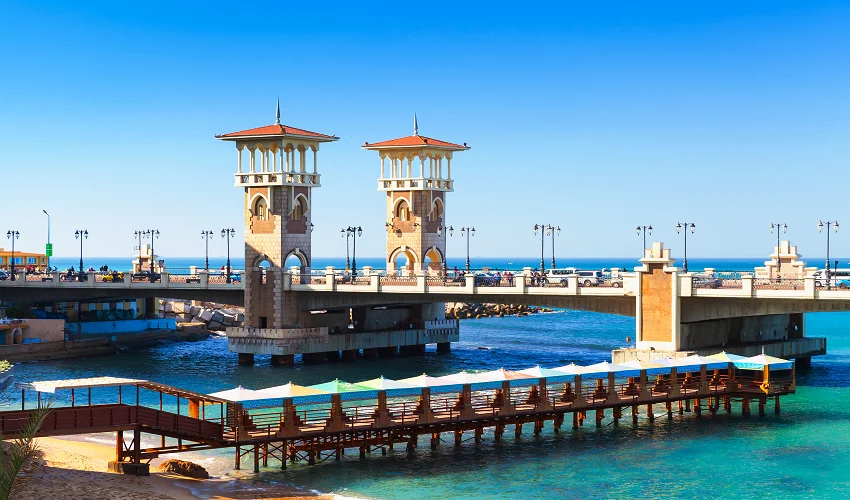Stanley-Brücke in Alexandria mit Zwillingstürmen im osmanischen Stil über türkisfarbenem Mittelmeer, davor Sonnenschirme