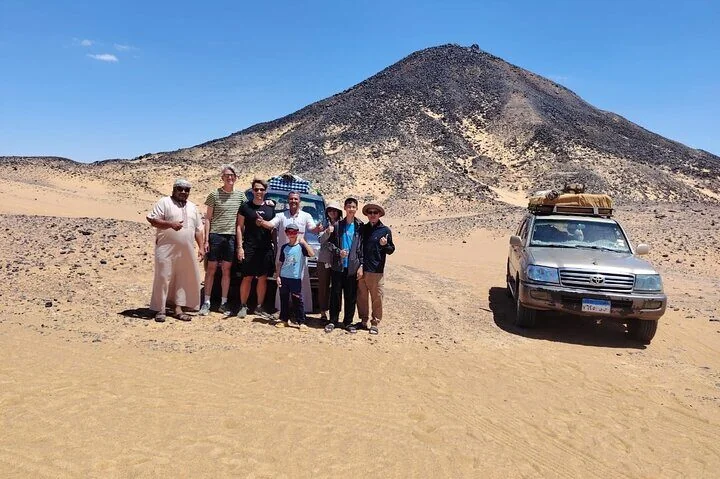 Einheimischer Guide und Touristen, auch Kinder, posieren vor einem Geländewagen und einem schwarzen Vulkankegel