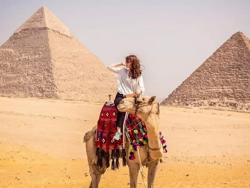 امرأة تركب جملًا بين هرمين من أهرامات الجيزة، ورمال الصحراء في المقدمة