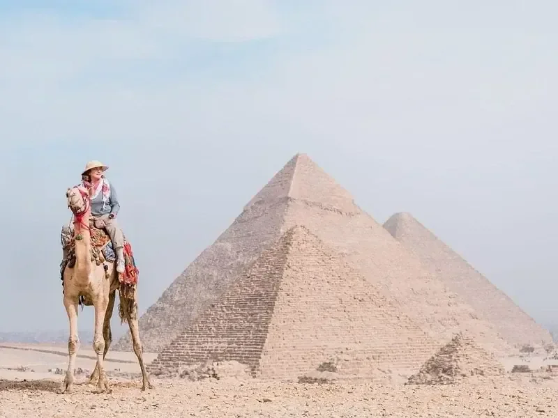 سائح جالس على جمل مصوَّر من الجانب قرب الهرم الأكبر في ضوء صحراوي ضبابي