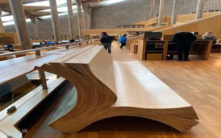 Interior of the Bibliotheca Alexandrina library showing a large wooden book sculpture and visitors at reading desks.