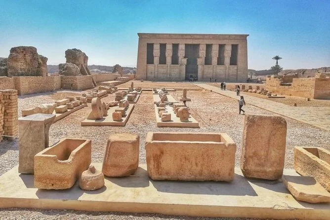 Ancient stone sarcophagi and carved fragments laid out in the temple courtyard in front of the Dendera Temple building.