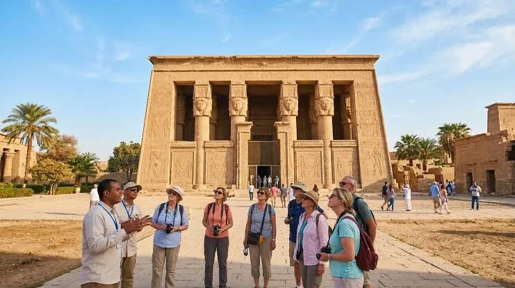 Tour guide speaking to a group of visitors in front of the Dendera Temple entrance with palm trees on either side.