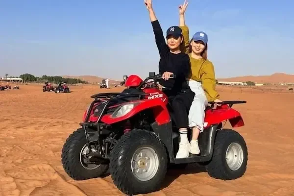 两位女士共乘一辆红色四轮摩托穿越沙漠，都举起手比出和平手势
