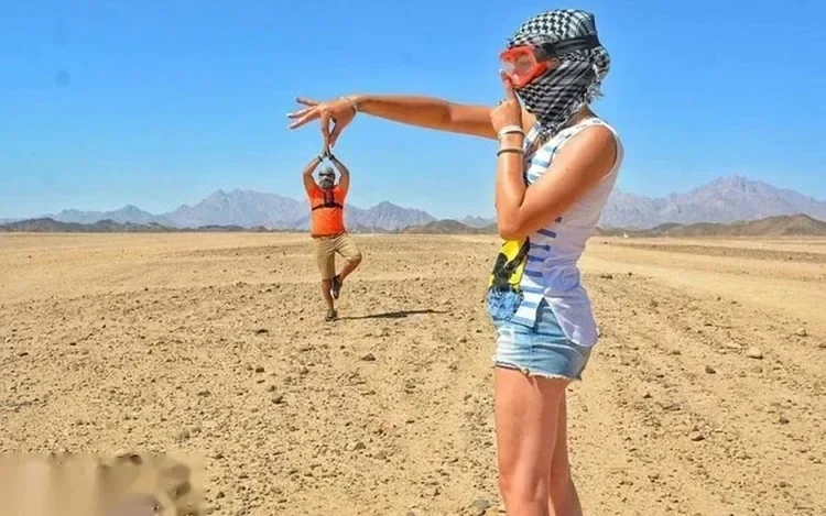 Snorkelmaszkot viselő nő a fején nézi, ahogy egy férfi ugrópózt vesz fel a sík sivatagban, a háttérben hegyekkel