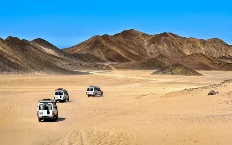Három fehér 4x4-es jármű halad konvojban egy nyílt sivatagi völgyön át, sötét hegyekkel körülvéve