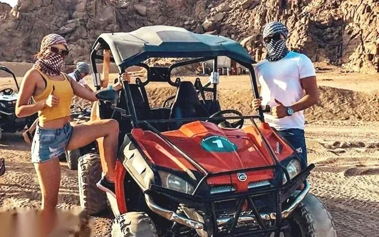 Man and woman giving thumbs up beside a red off-road buggy parked in front of rocky desert mountains.