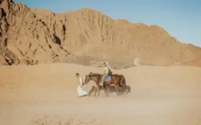 Two people on horseback leading a third horse across open sandy desert with mountains in the background.