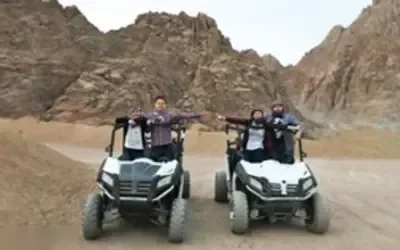 Two off-road buggies with riders parked side by side in front of jagged desert mountains.