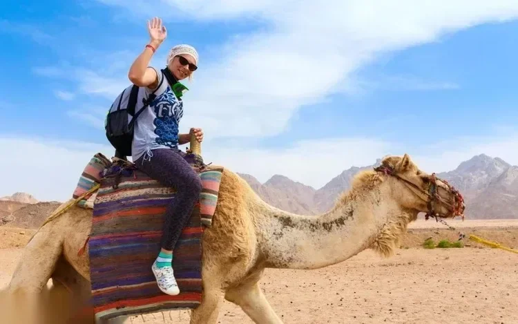 Woman riding a camel through the desert, waving at the camera, with mountains in the distance.