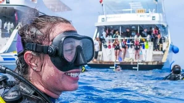 غواص مبتسم بقناع، وخلفه قارب غوص كبير وغواصون آخرون