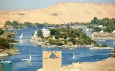 منظر واسع للنيل في أسوان مع فلايك تبحر مارّة بصخور جرانيتية ومبنى مقبّب على ضفة النهر