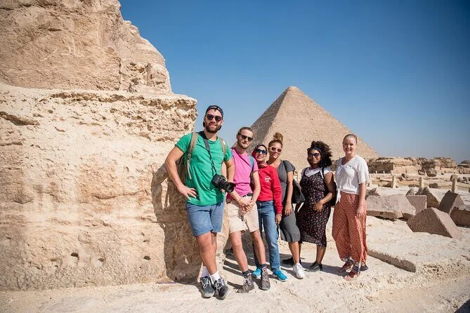 A group of six tourists smiling beside a rock outcrop with the Great Pyramid visible behind them.